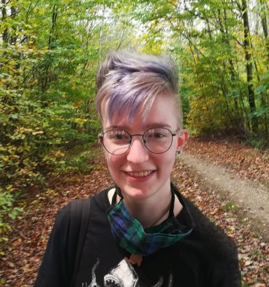 Foto von Koi Katha Blaeser. Eine Person mit kurzen blond-blauen Haaren, einer dunklen Brille, mit Maske um den Hals und schwarzen T-Shirt. Im Hintergrund sind Bäume zu sehen und ein Weg mit sehr vielen heruntergefallenen Blättern.